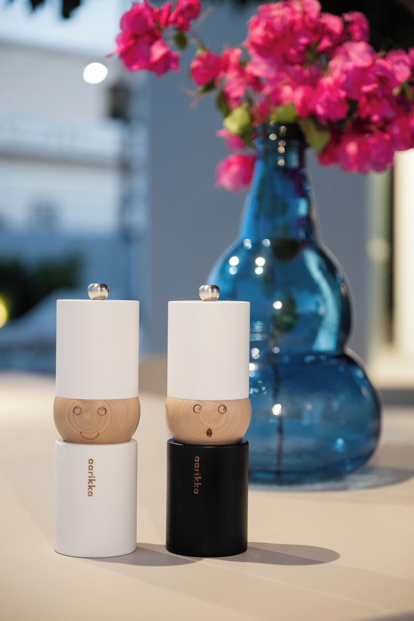 Two candles with wooden bases and a black candle on a table, with a blue vase with pink flowers in the background.
