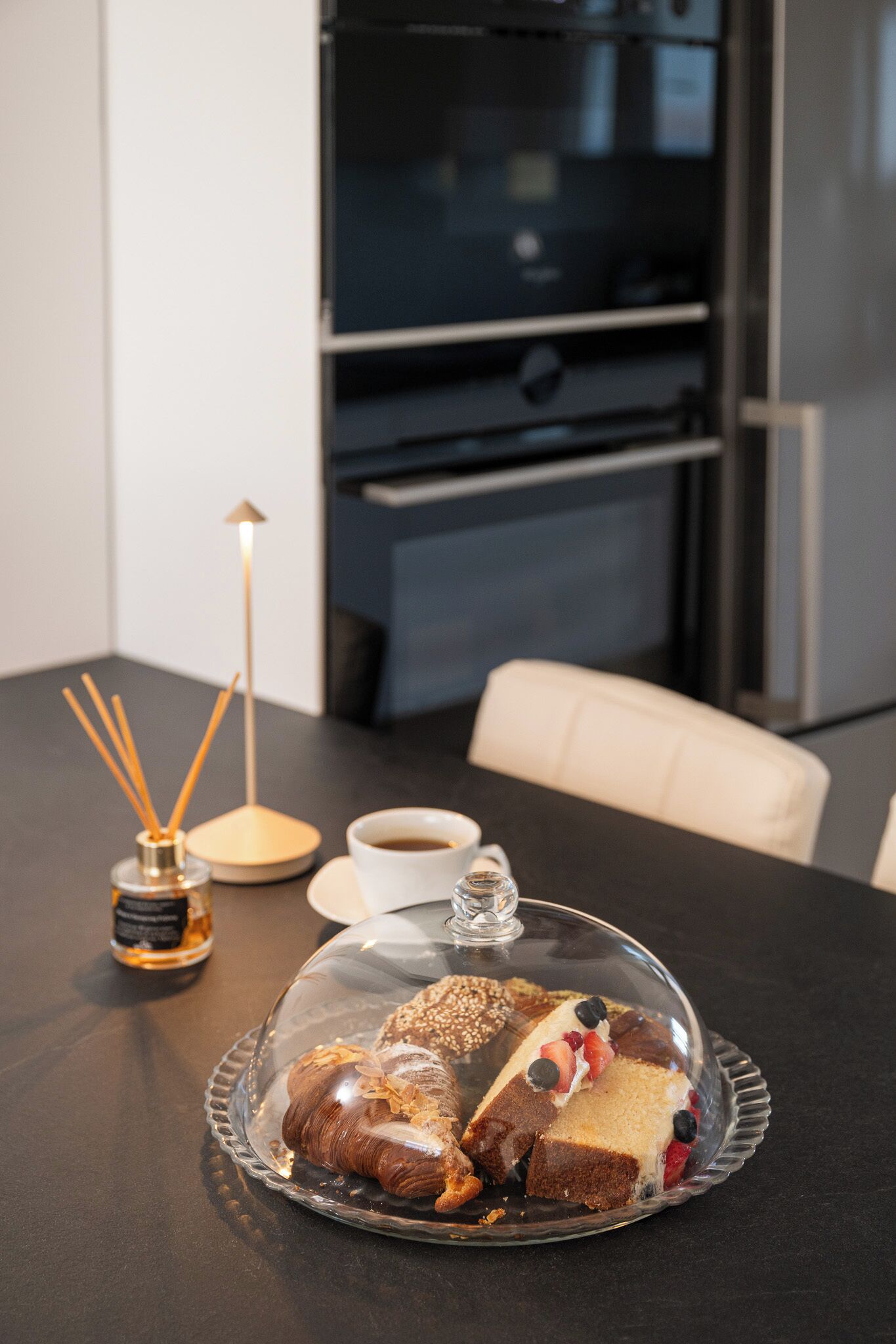 Una mesa de comedor con una campana de cristal sobre un postre, una taza de café y una botella de perfume.