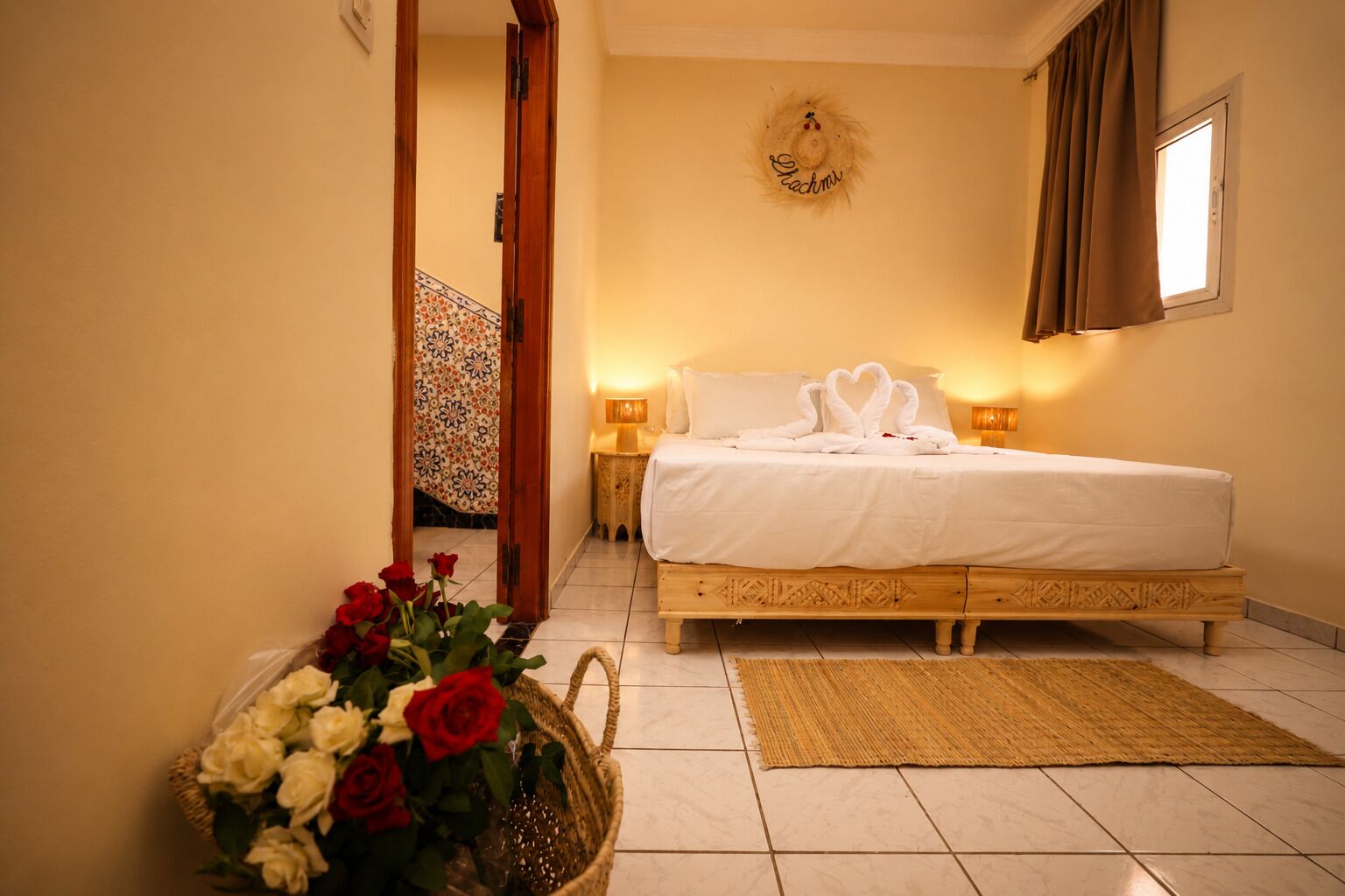 A hotel room with a bed, bedside lamps, a decorative wall hanging, a woven basket with flowers, and a patterned rug.