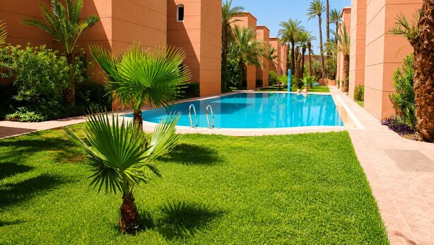 A swimming pool surrounded by palm trees and greenery, with buildings in the background. - Riad Kamin And Pool (Marrakech)