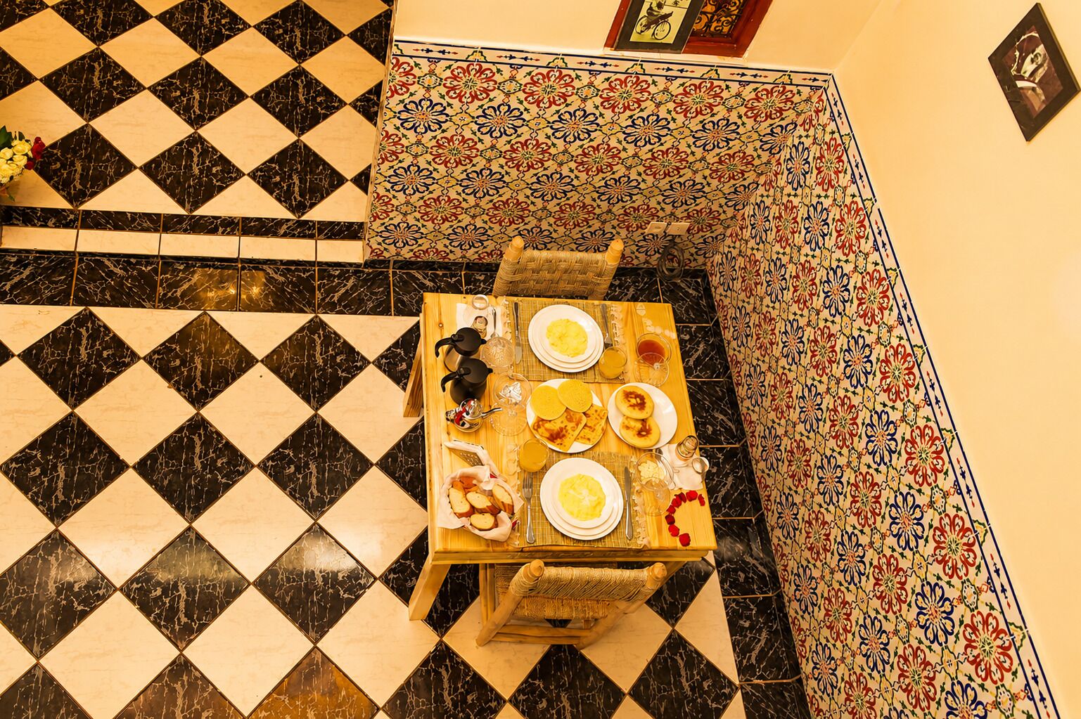 A dining table with plates, glasses, and a woven basket, set on a black and white checkered floor with a patterned wall in the background.