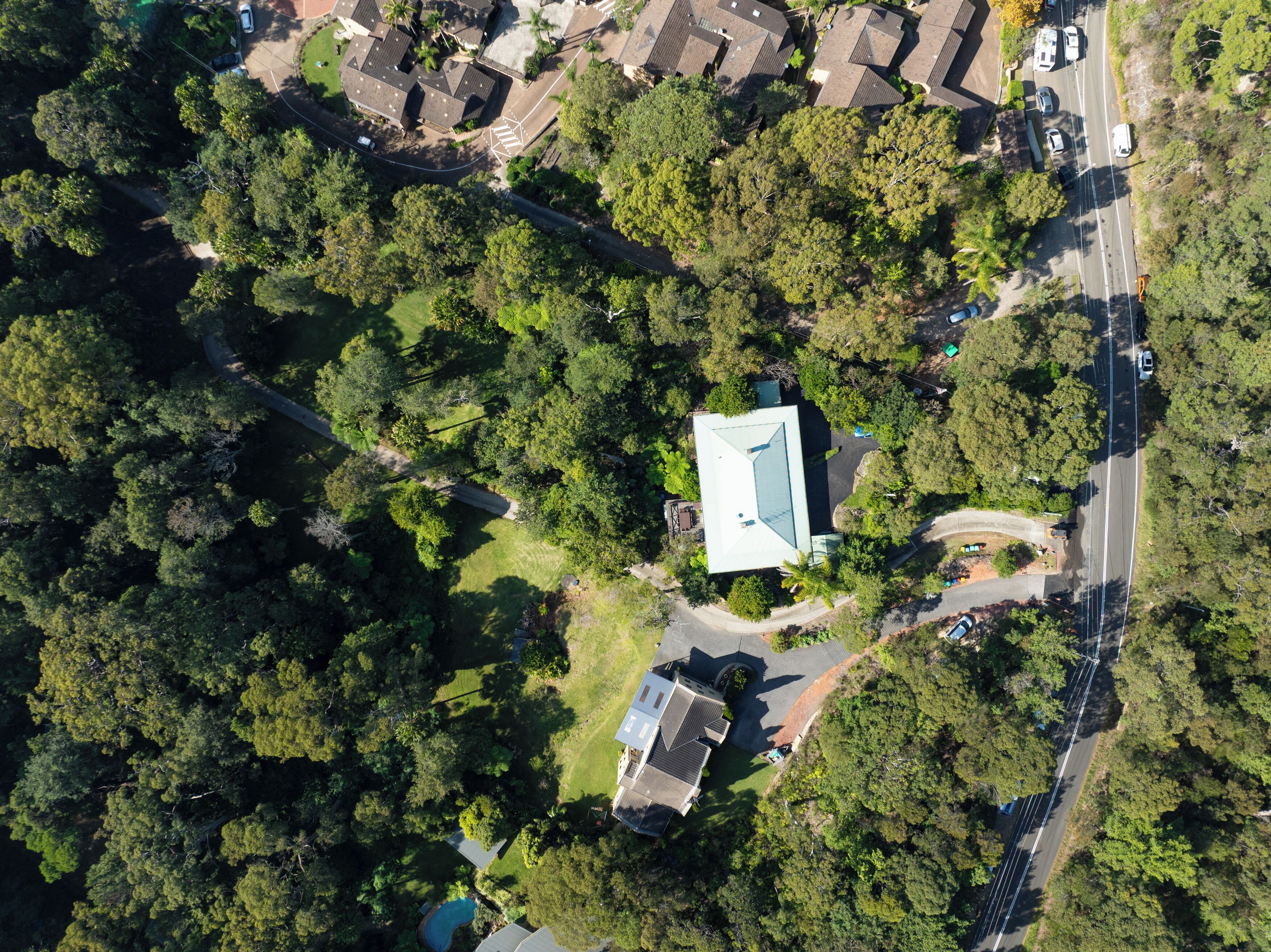 An aerial view of a residential area with a house featuring a sloped roof, surrounded by dense greenery and a road with vehicles.