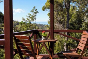 Une terrasse en bois avec deux chaises et une petite table, offrant une vue sur un paysage boisé et la mer.