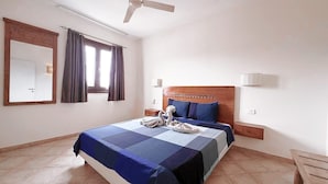 A hotel room with a bed, a ceiling fan, a window with curtains, a bedside table, and a nightstand. - HostySpot - Tropical Resort, 1-Bedroom Apartment (Santa Maria)