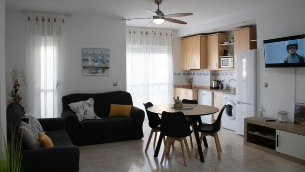 Una sala de estar moderna con un sofá, una mesa de comedor con sillas, un ventilador de techo y una cocina con armarios y electrodomésticos.