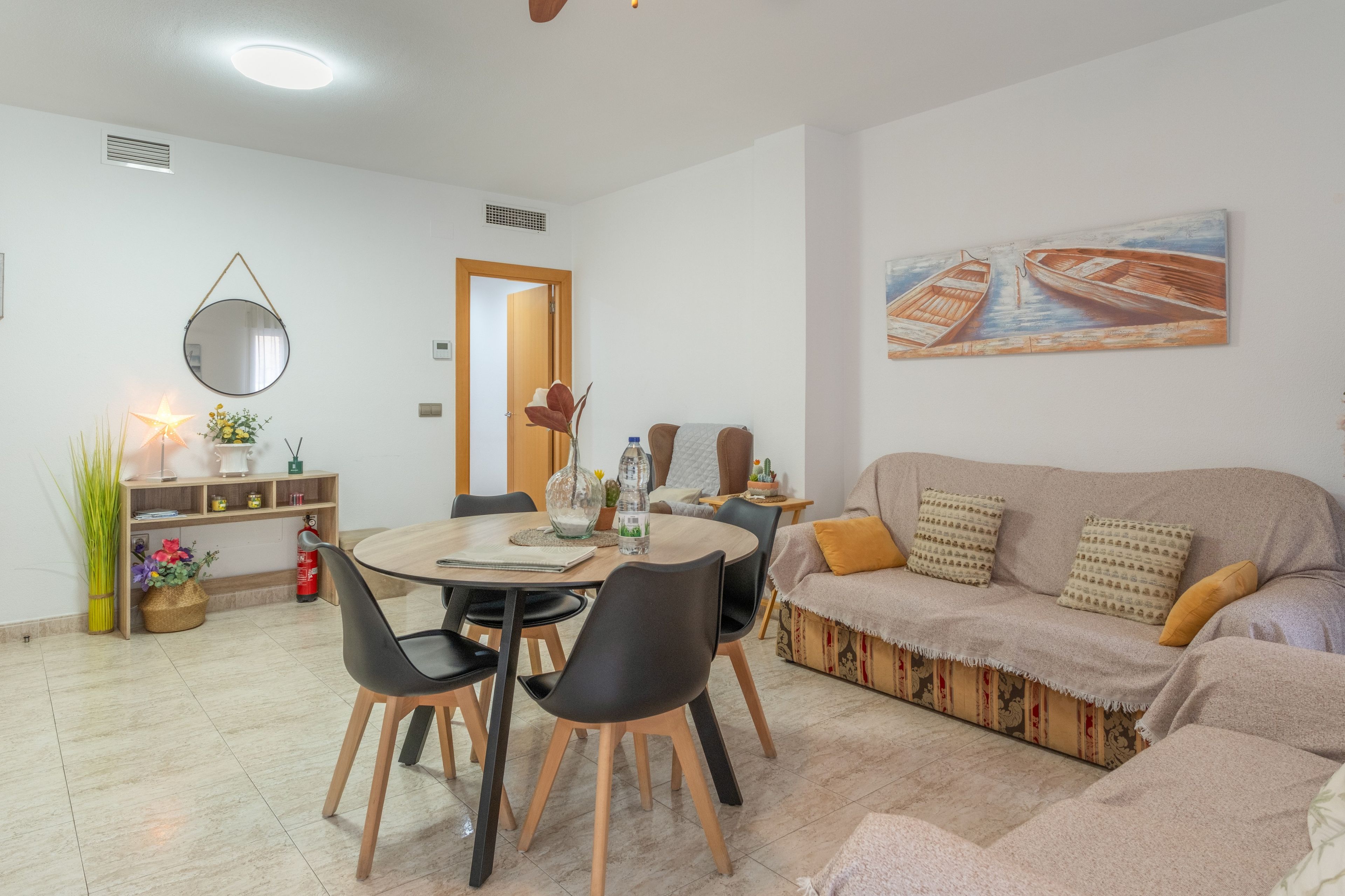 A living room with a sofa, a dining table with chairs, a coffee table, a painting on the wall, and a door leading to another room.