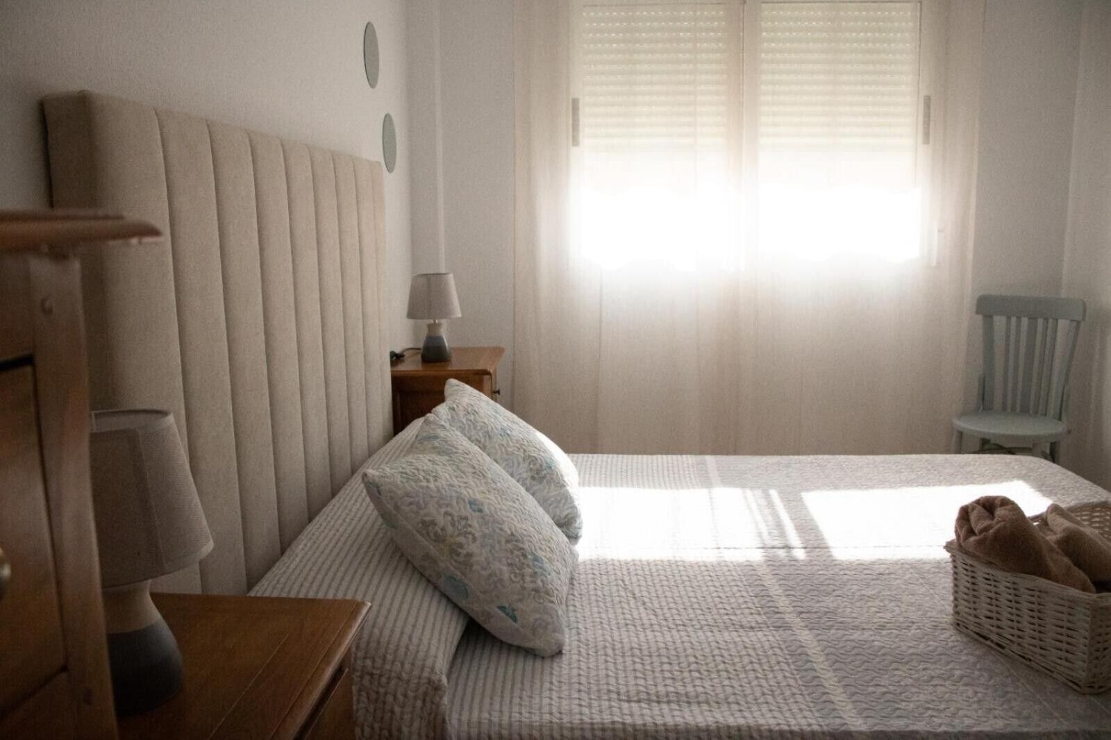 Een slaapkamer met een bed, kussens, een nachtkastje, een lamp, een stoel en een raam met jaloezieën.