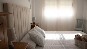 Een slaapkamer met een bed, kussens, een nachtkastje, een lamp, een stoel en een raam met jaloezieën.