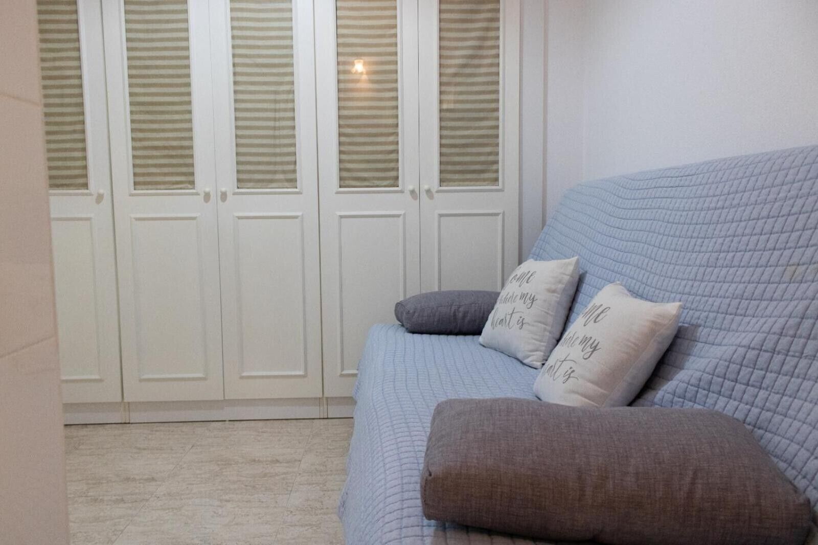 A room with a blue sofa, two pillows, a light fixture, and a set of white doors with horizontal blinds.