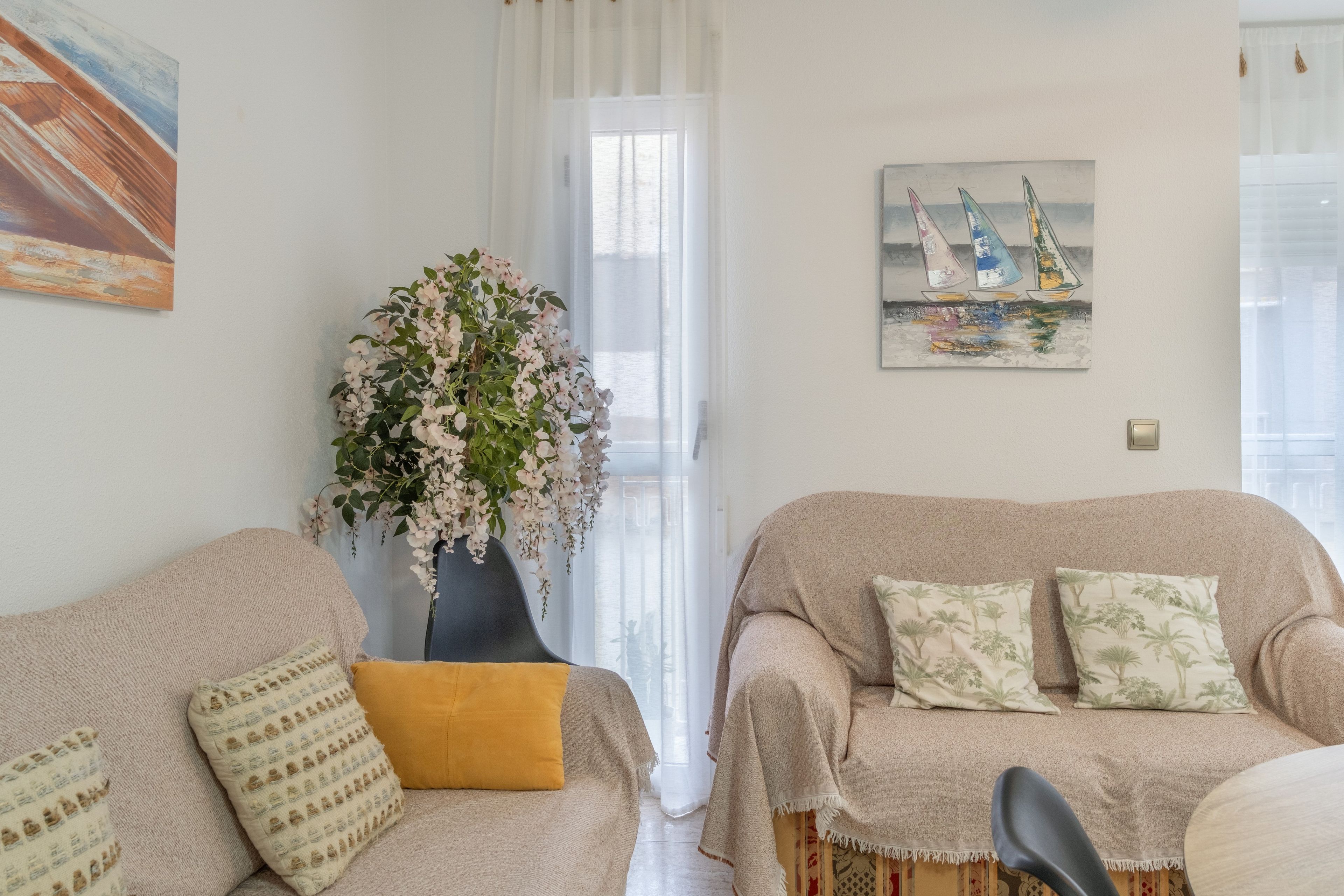 A living room with a sofa, a chair, a vase with flowers, a painting of sailboats, and a window with curtains.