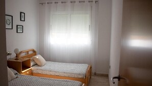 Un dormitorio con dos camas individuales, una mesita de noche con lámpara, una ventana con persianas y una puerta.