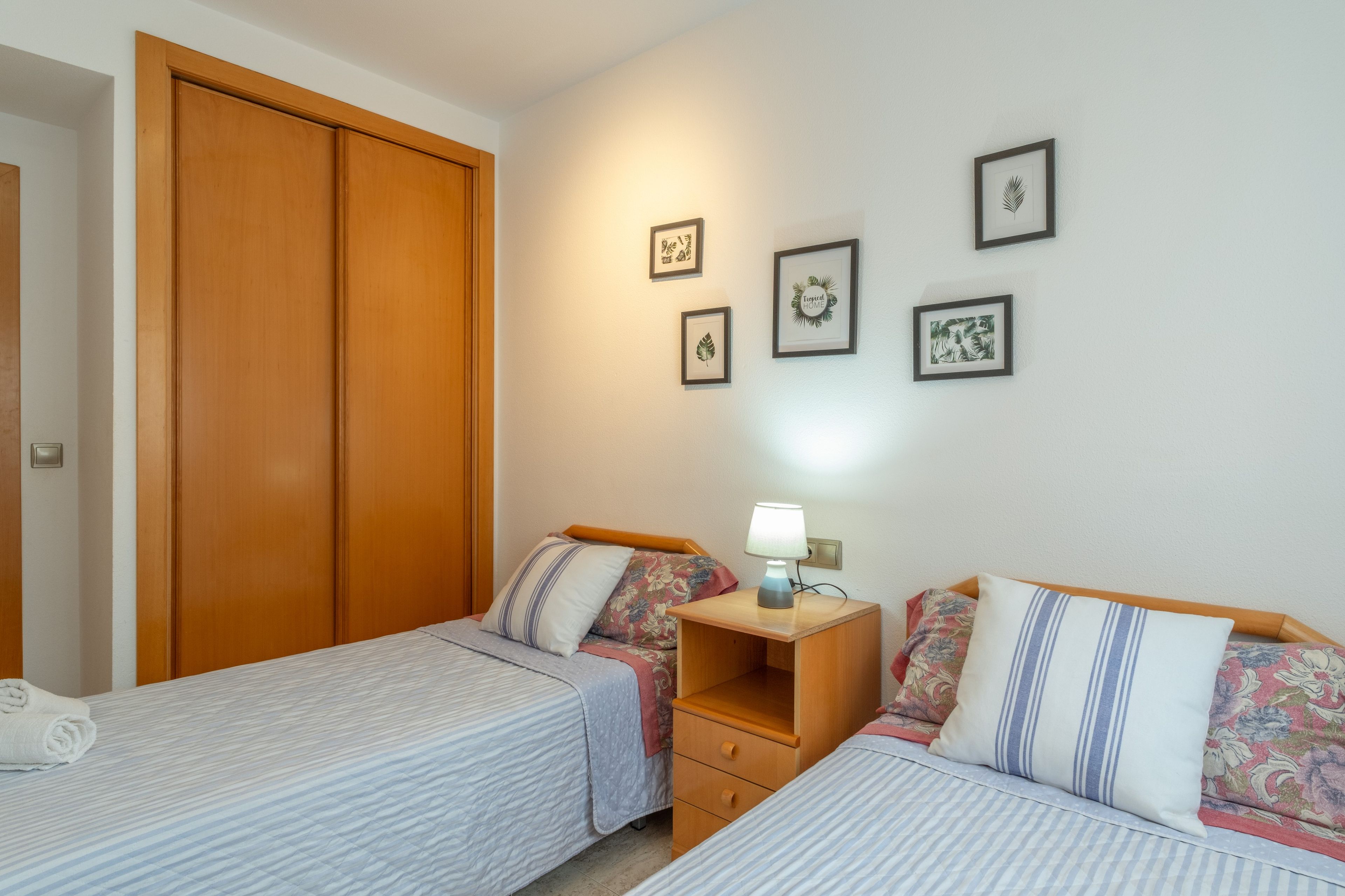 Un dormitorio con dos camas individuales, un armario de madera, una mesita de noche con lámpara y una obra de arte en la pared.