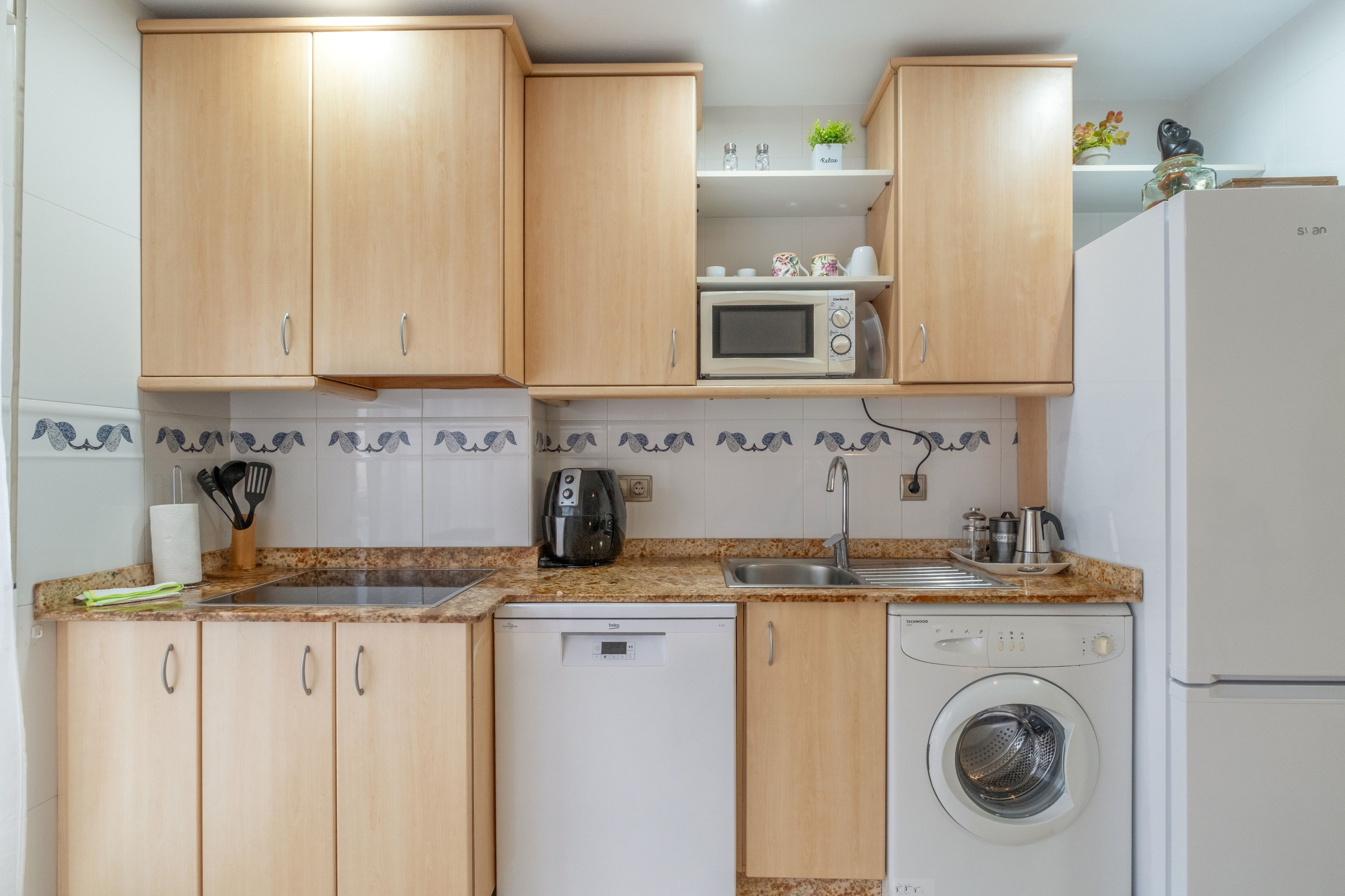 A compact kitchen with wooden cabinets, a microwave, a coffee maker, a sink, and a washing machine.
