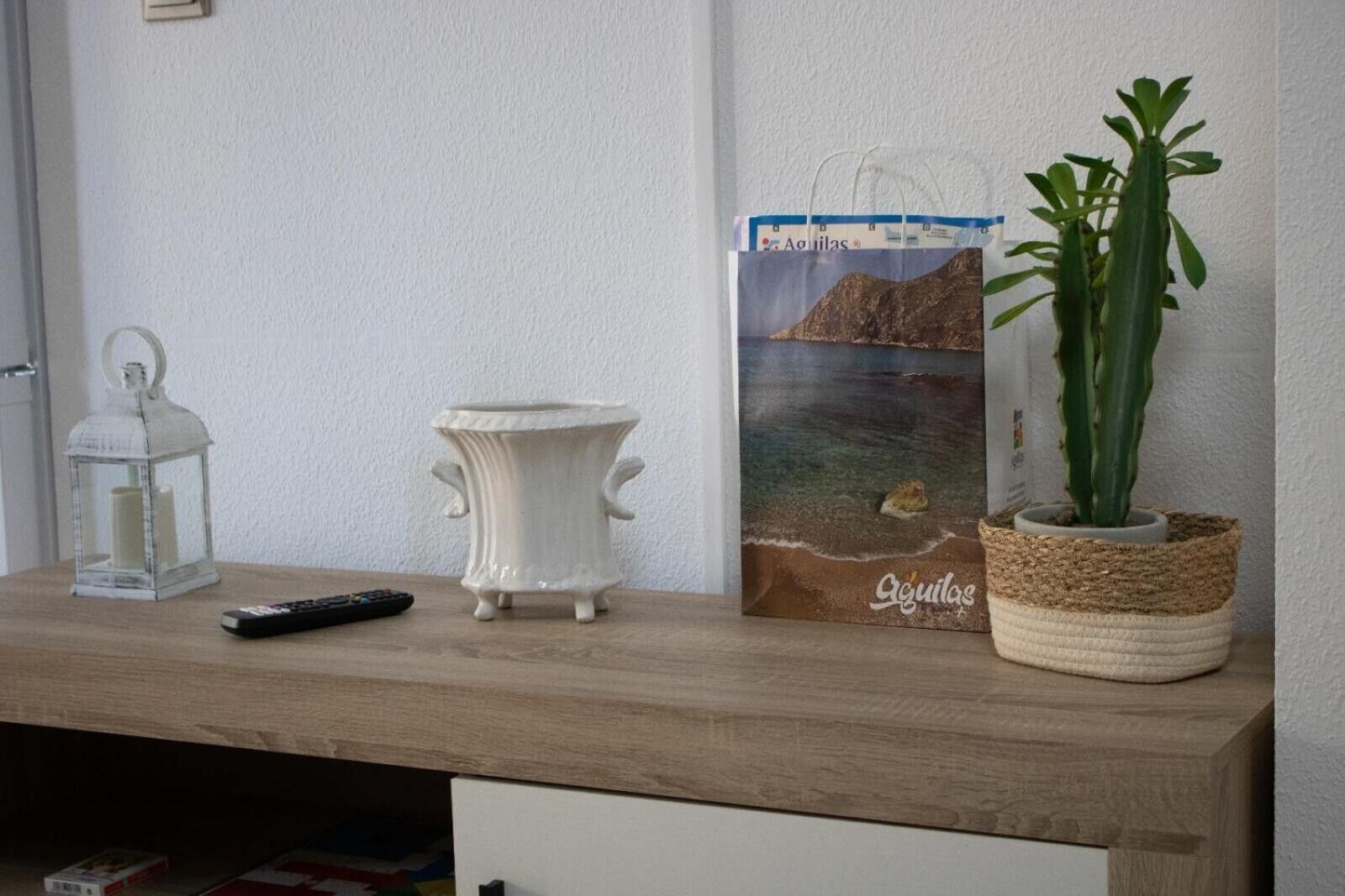Een houten console tafel met een afstandsbediening, een witte vaas, een lijst met foto en een plant in pot.