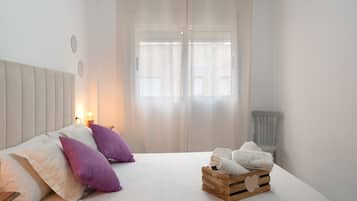 A bedroom with a bed, pillows, a bedside table, a nightstand, a window with curtains, and a chair.