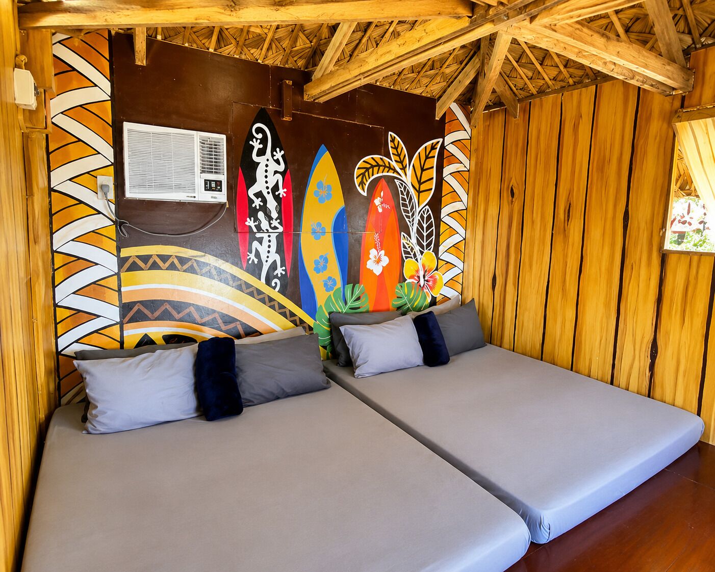 A room with two beds, a wooden ceiling, and a wall decorated with colorful surfboard designs.