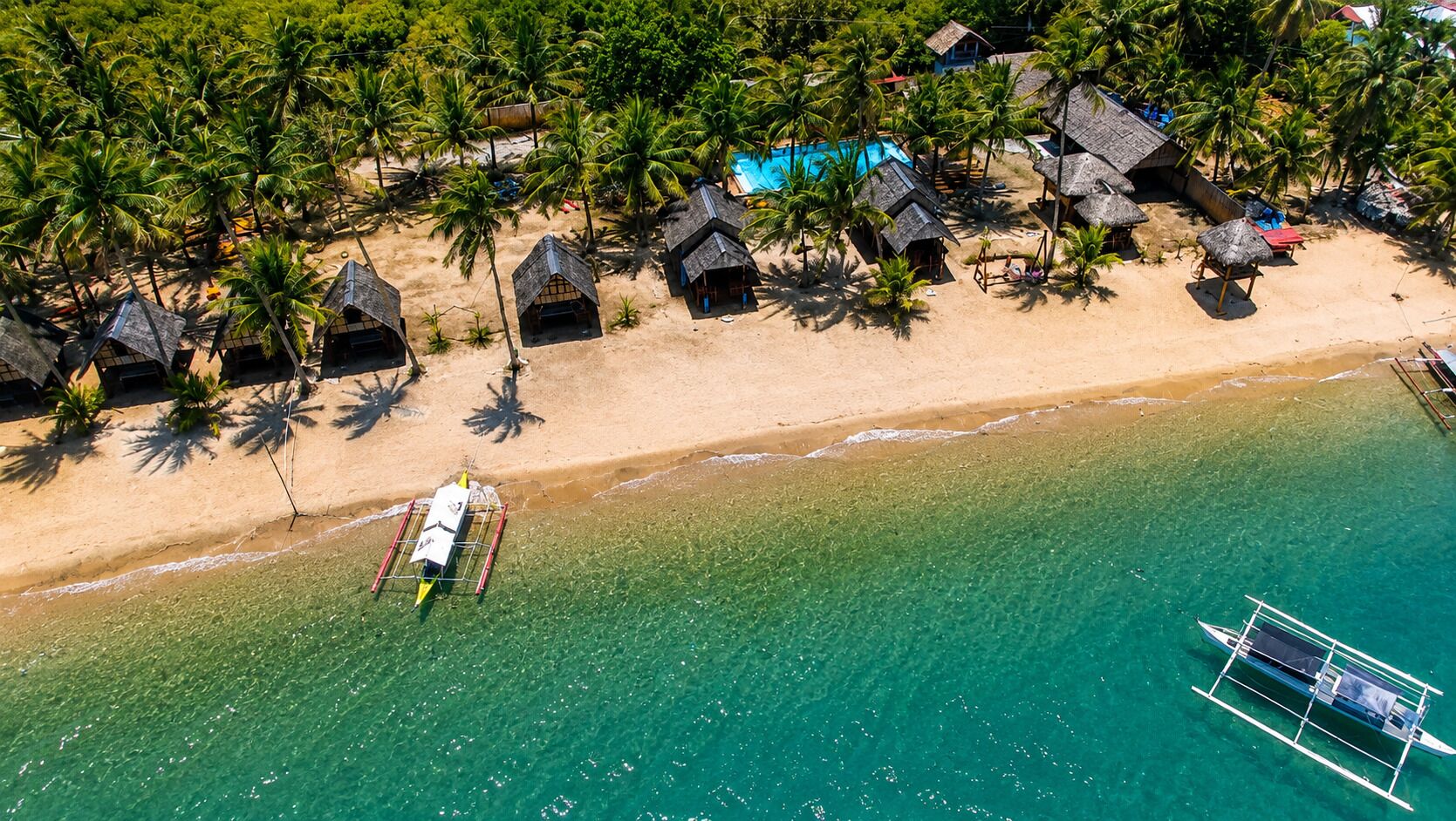 An aerial view of a beach with thatched-roof huts, clear turquoise water, and a boat docked near the shore.