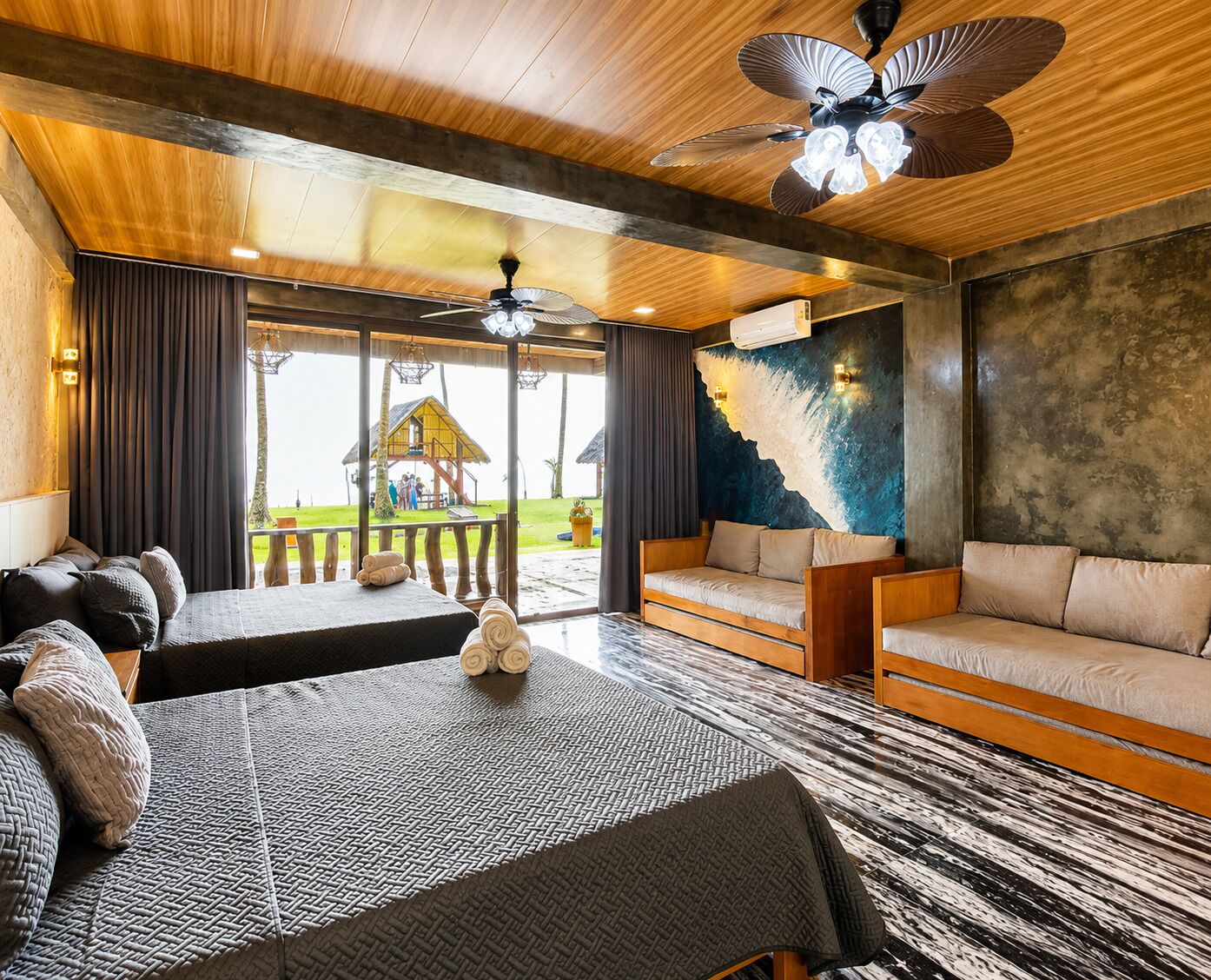 A modern hotel room with a large bed, a sofa, and a dining table. There is a view of a pool and a thatched-roof structure outside.