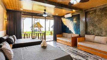 A modern hotel room with a large bed, a sofa, and a dining table. There is a view of a pool and a thatched-roof structure outside.
