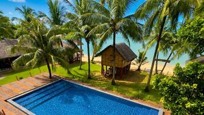 Piscina rectangular con una terraza de madera alrededor, frente a un telón de fondo de palmeras y una playa.