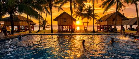 A swimming pool with palm trees and beach huts, surrounded by a sunset view.