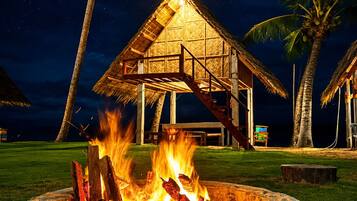 A stone fire pit with a thatched-roof structure above it, surrounded by palm trees and a night sky.