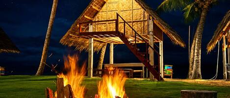 A stone fire pit with a thatched-roof structure above it, surrounded by palm trees and a night sky.