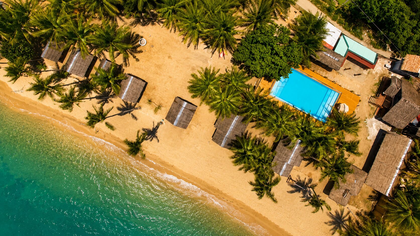 Vista aérea de un complejo turístico frente a la playa con piscina, palmeras y cabañas.
