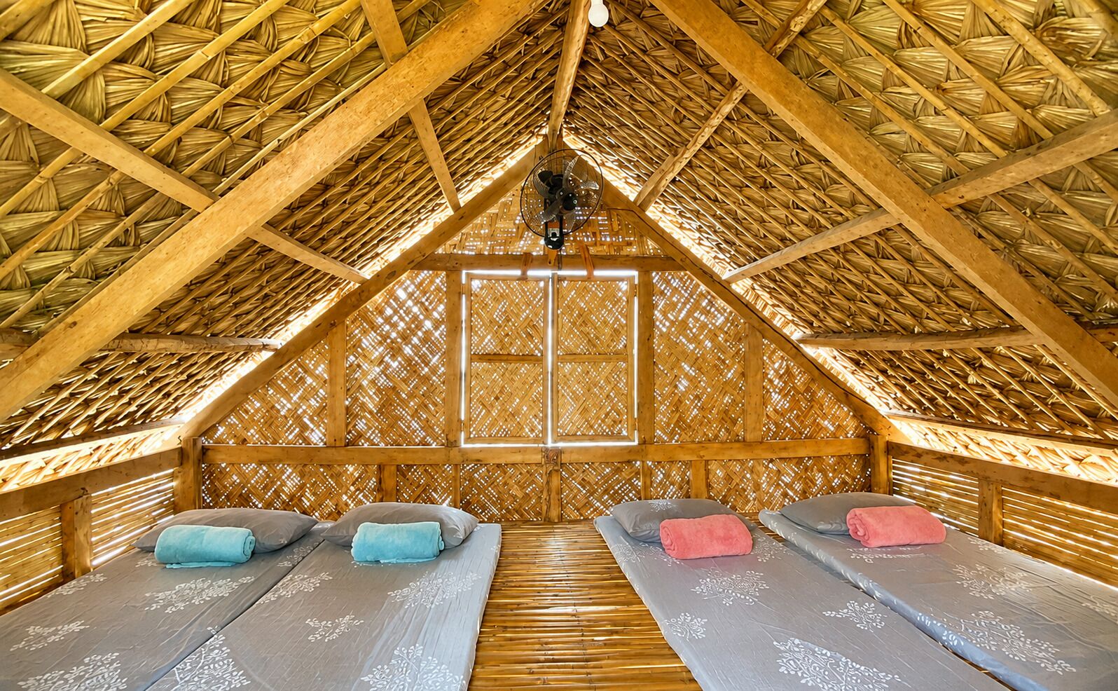A room with a thatched roof, two beds with pillows, and a fan.