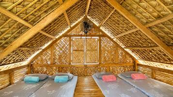 A room with a thatched roof, two beds with pillows, and a fan.