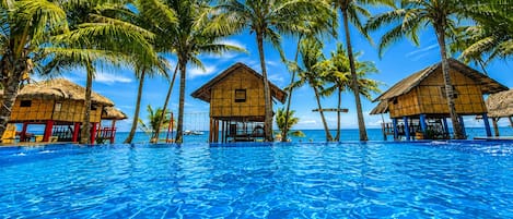 Une piscine d’un bleu éclatant, entourée de bungalows sur pilotis aux toits de chaume, de palmiers et offrant une vue sur l’océan.