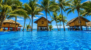 A clear blue pool surrounded by overwater bungalows with thatched roofs, palm trees, and a view of the ocean. - Calilayan Cove Beach Resort (Unisan)