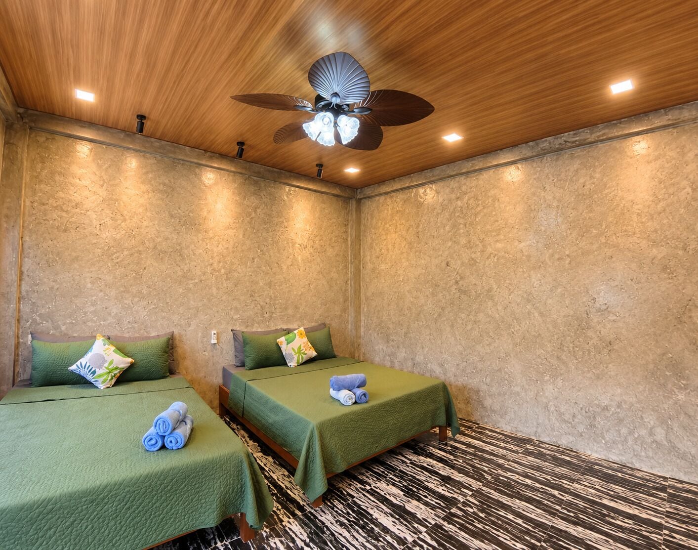 A room with two beds, each with a green bedspread and a pillow, a ceiling fan, and a textured wall.