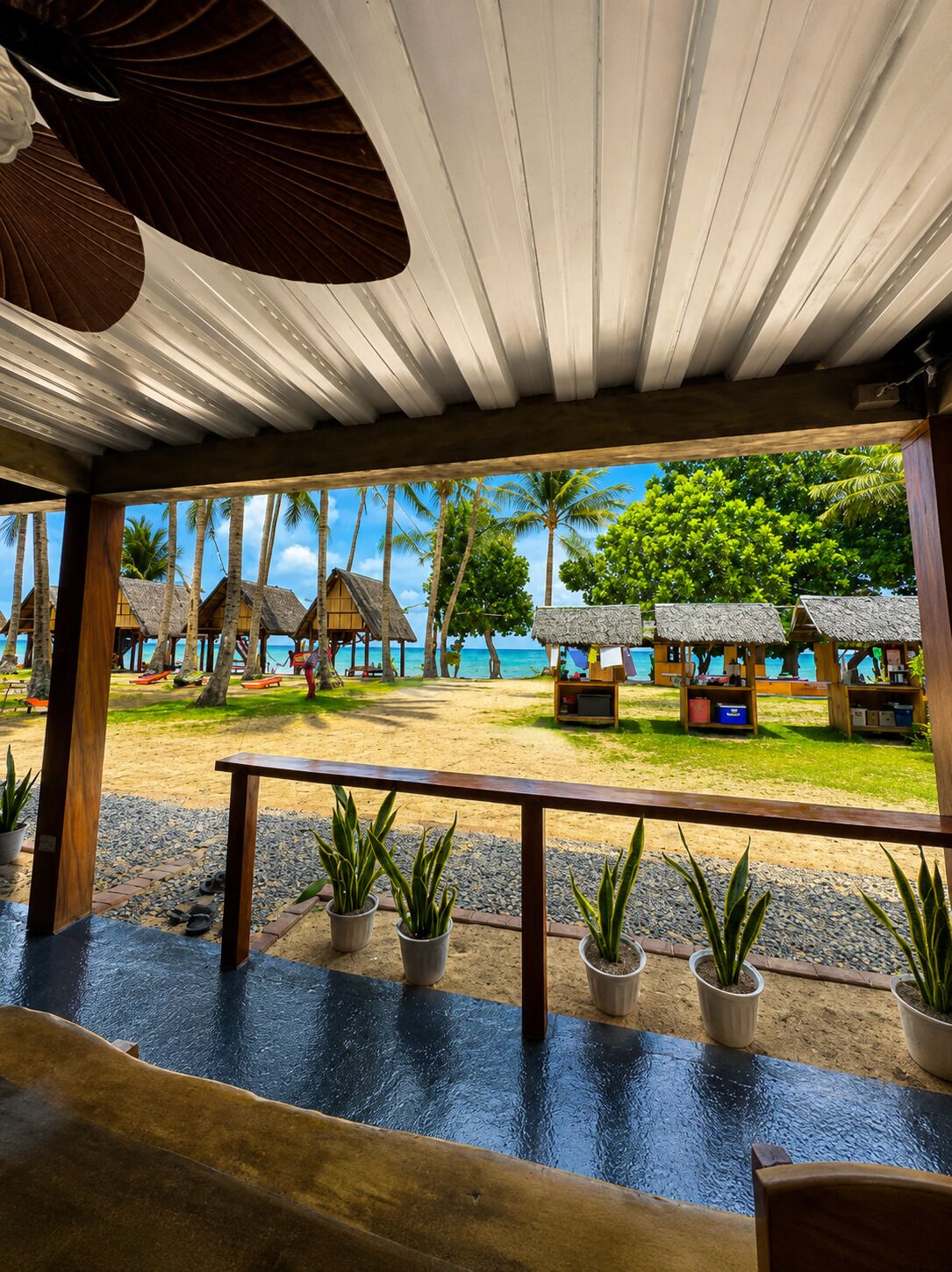 A covered outdoor area with a view of a beach and thatched-roof structures.