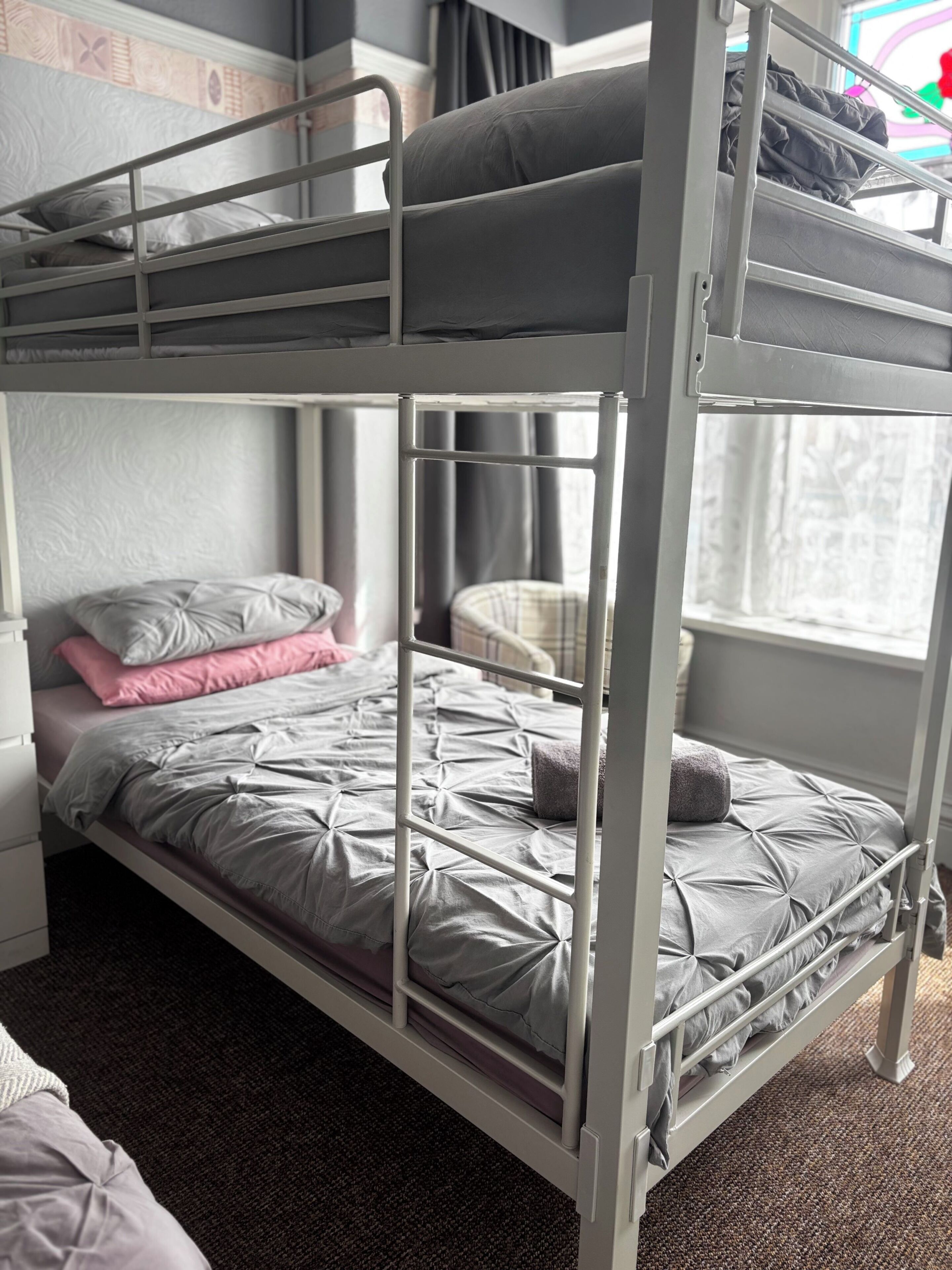 Un lit superposé avec un matelas et des oreillers gris, une petite chaise et une fenêtre avec des rideaux.