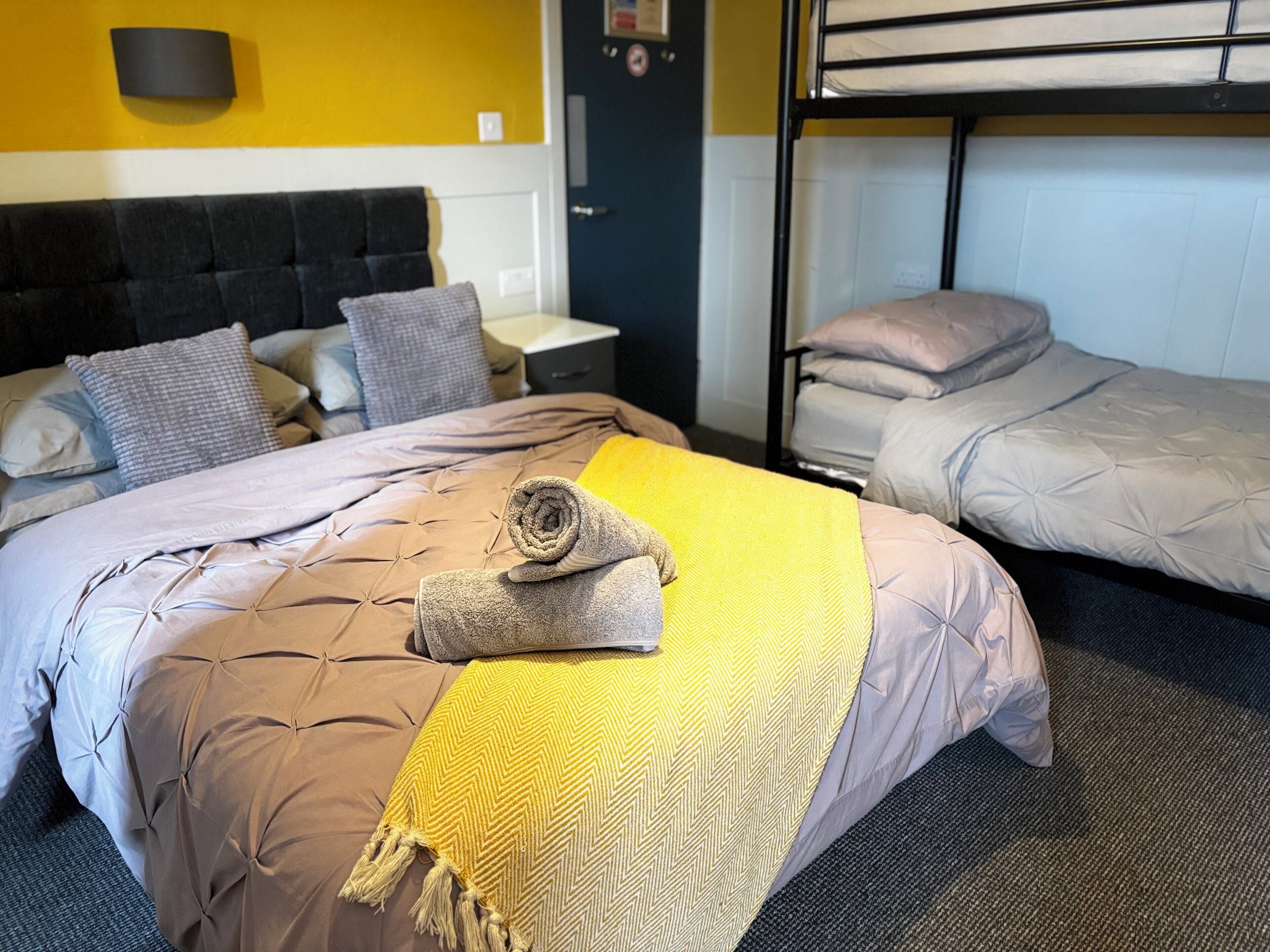 Quarto de hotel com cama, mesa de cabeceira, beliche e luminária fixada na parede.