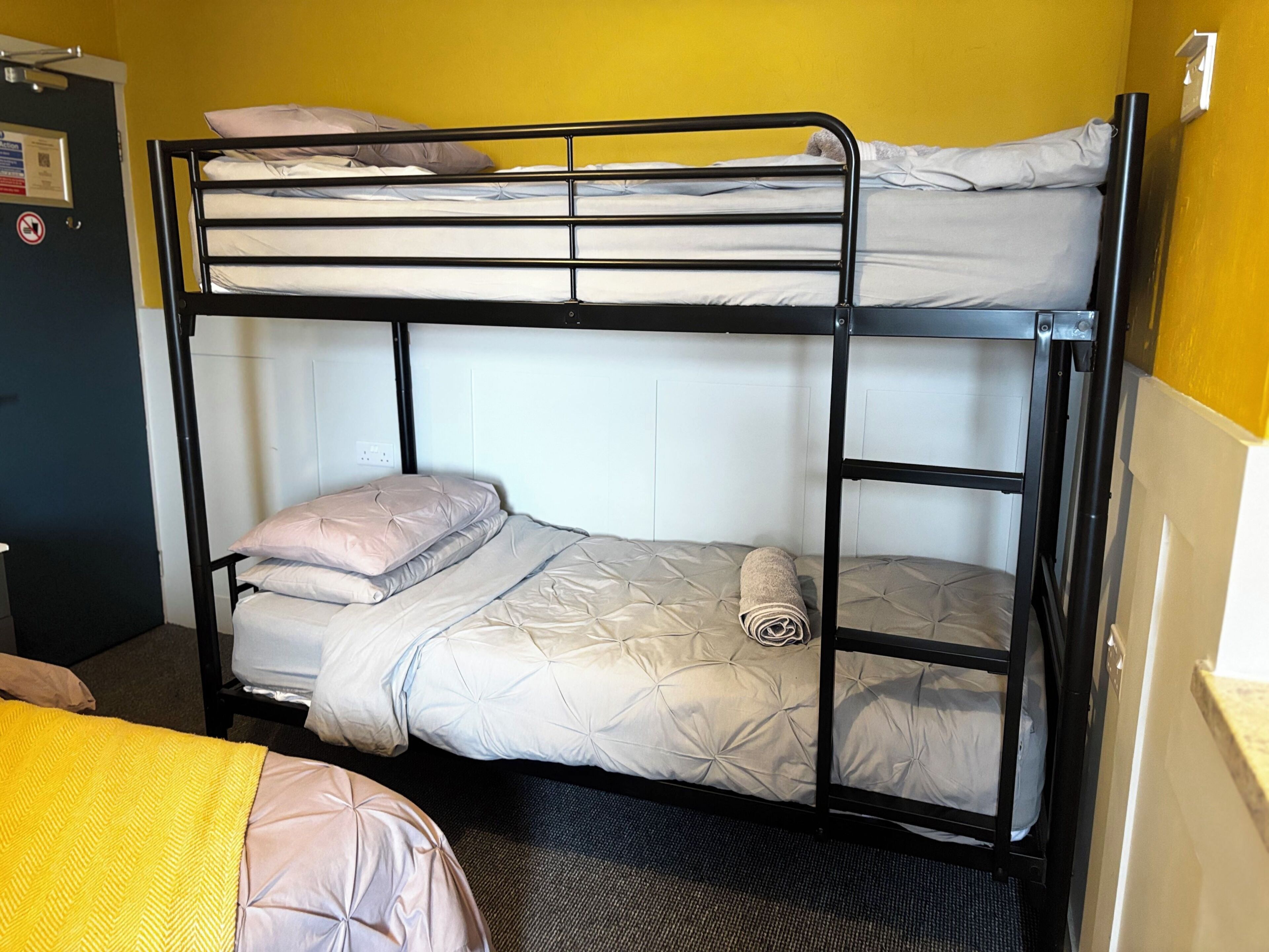 Una cama litera con colchón y almohadas, una puerta con un cartel, y una pared con interruptor de luz.