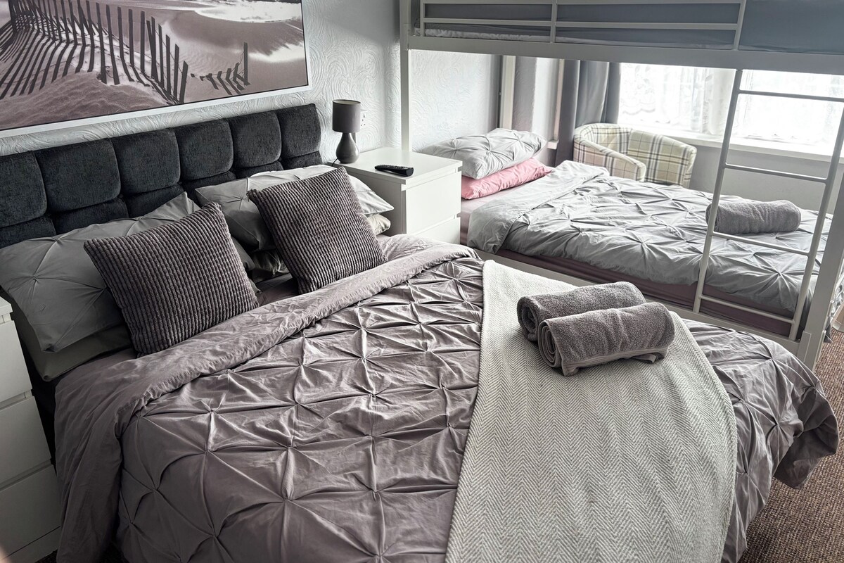A room with a bunk bed, a single bed with a headboard, a bedside table, and a wall-mounted lamp.