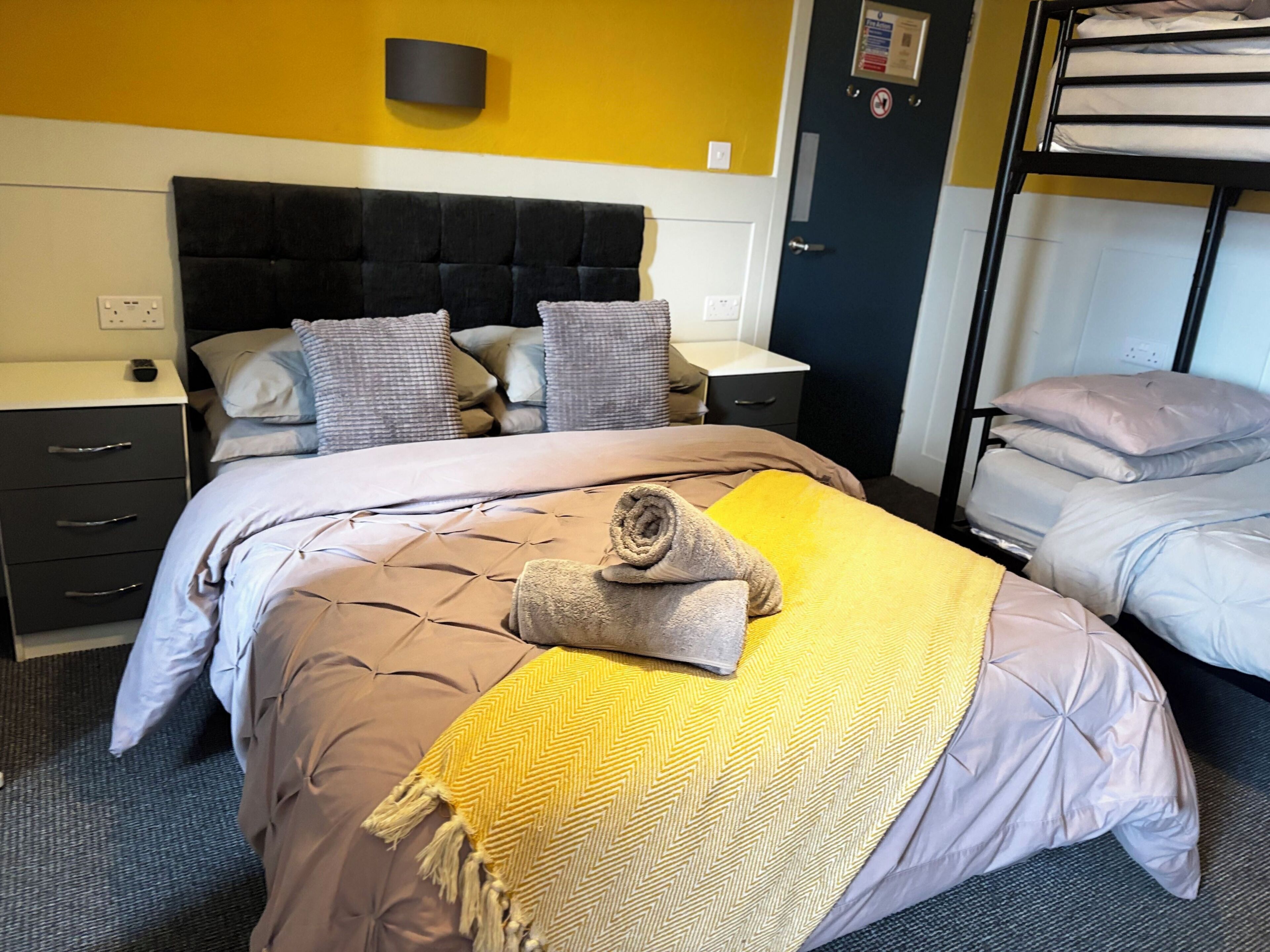 A hotel room with a bed, a nightstand, a dresser, a bunk bed, and a wall-mounted light fixture.