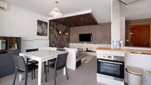 A modern kitchen with a built-in oven, a dining area with a table and chairs, a TV mounted on the wall, and a wooden ceiling.