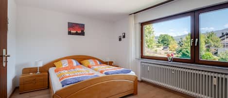 Un dormitorio con cama de madera, una mesita de noche, un ventanal con vistas a la vegetación y un radiador.