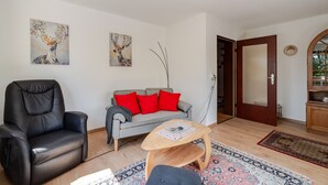 Ein Wohnzimmer mit Ledersessel, grauem Sofa mit roten Kissen, Couchtisch aus Holz und zwei an der Wand hängenden Bildern.