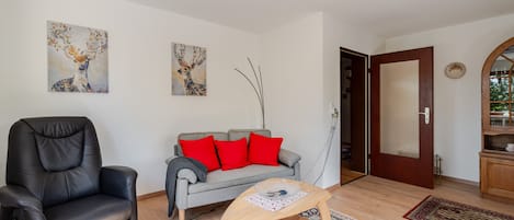 A living room with a leather chair, grey sofa with red cushions, a wooden coffee table, and two wall-mounted paintings.