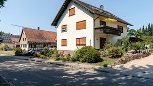 Ein zweistöckiges Haus mit Balkon, einem geparkten Auto davor und einem Garten mit verschiedenen Pflanzen.
