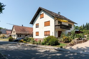 A two-story house with a balcony, a car parked in front, and a garden with various plants. - Quiet apartment “Haus Schwarzwaldblick” with mountain view, balcony, and Wi-Fi by the stream (Ottersweier)
