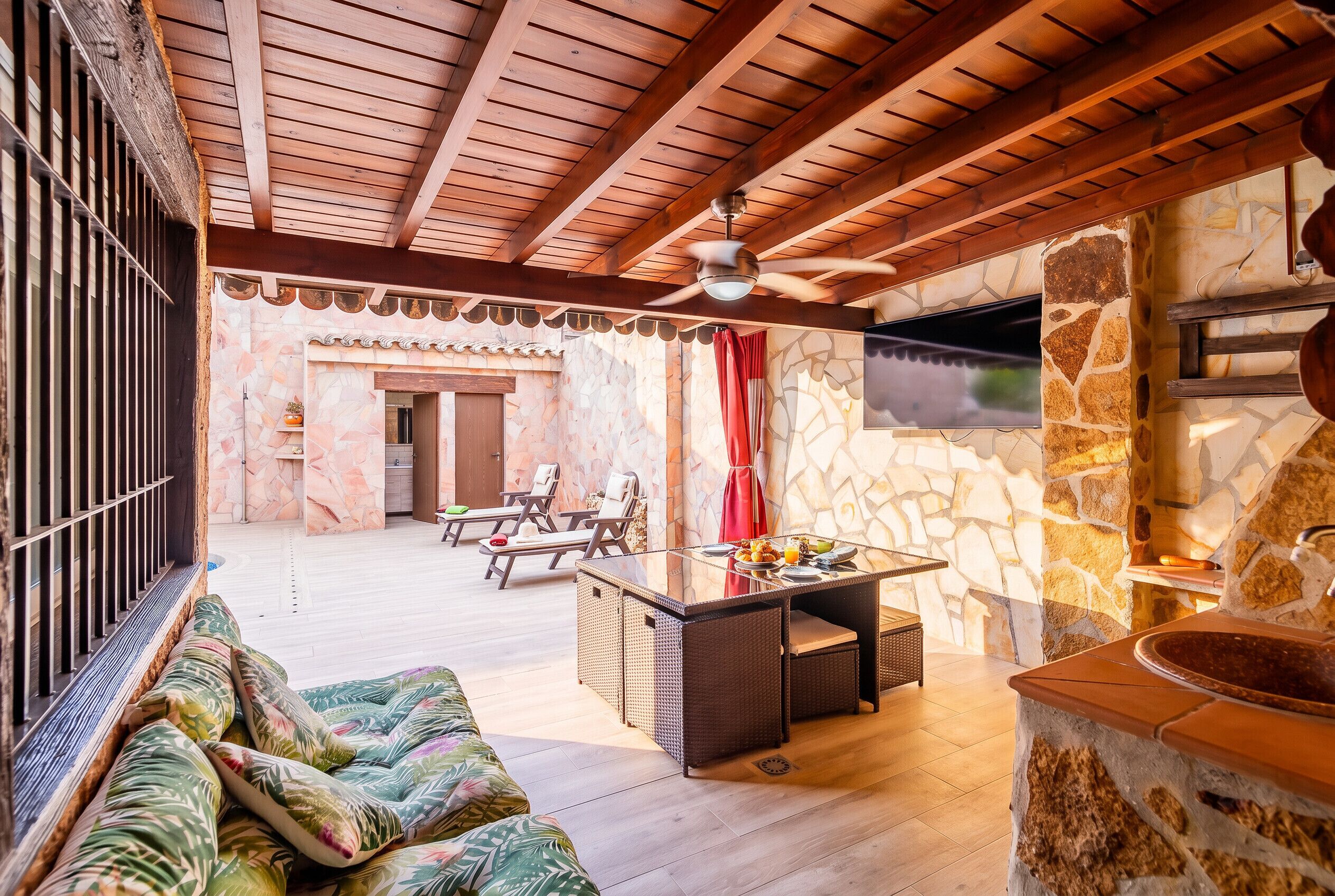 Un espacio al aire libre iluminado por el sol, con pared de piedra, techo de madera, mesa de billar y sillones de descanso.