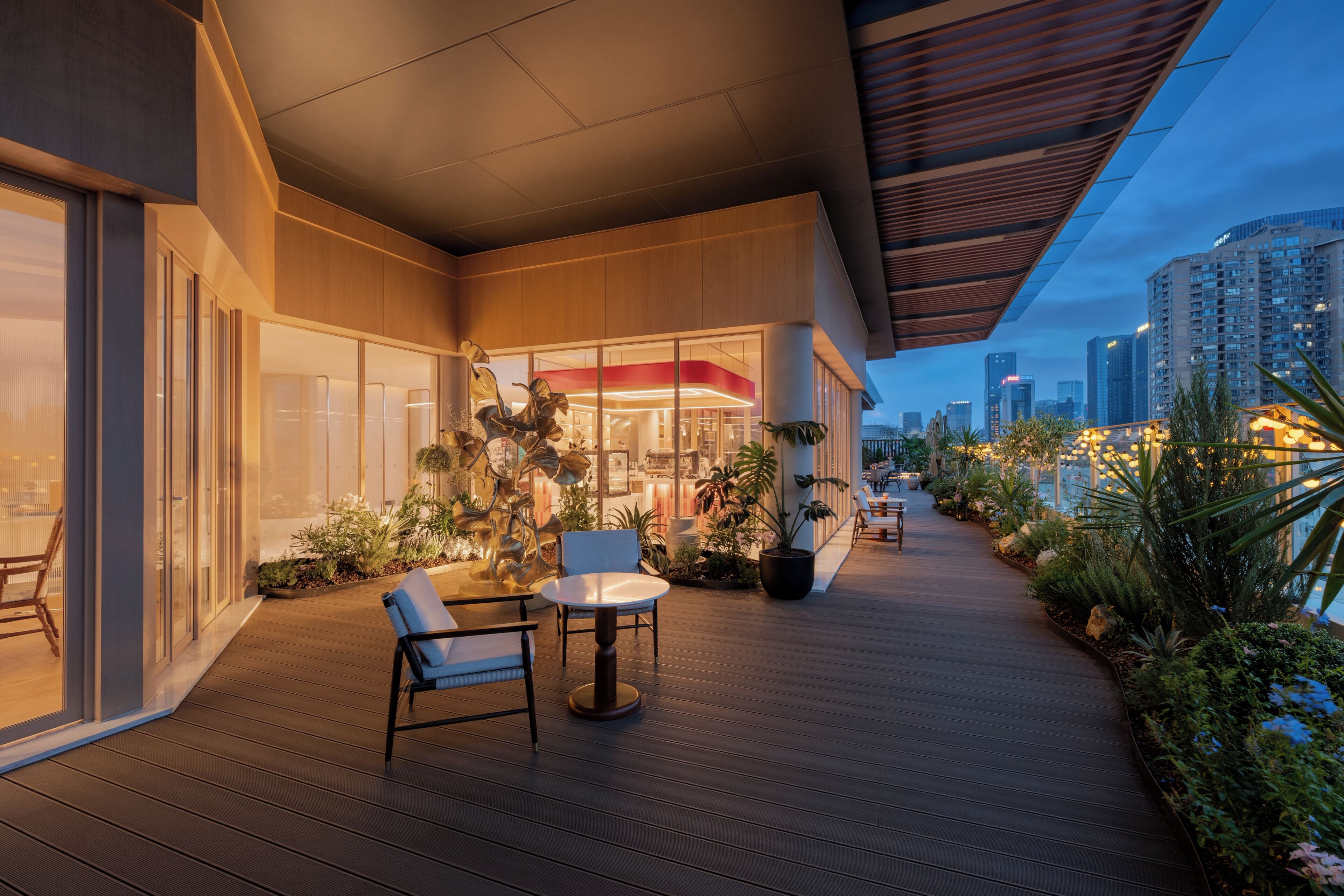 Un espacio exterior moderno con terraza de madera, mobiliario y un edificio con fachada de vidrio.