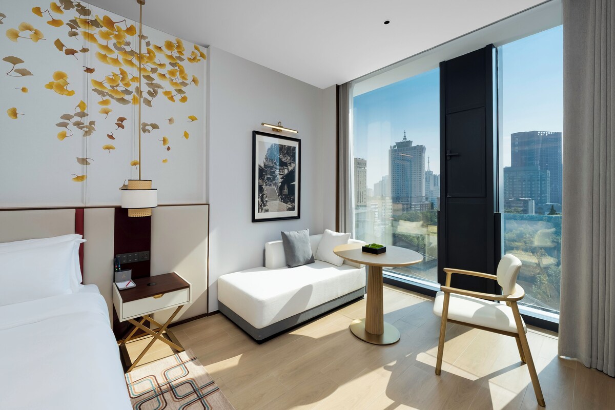 A modern hotel room with a large window, a sofa, a small round table, and a bed.