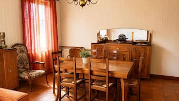 Uma sala de jantar com mesa e cadeiras de madeira, um lustre, uma cortina vermelha e um armário com objetos decorativos.