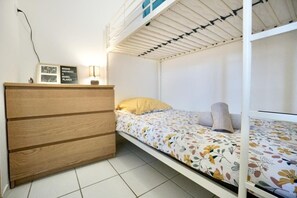 A single bed with a floral bedspread, a wooden dresser, and a wall-mounted shelf with framed pictures. - Swimming pool with a 100-meter cable winch, located 100 meters from the beach* (Guidel)